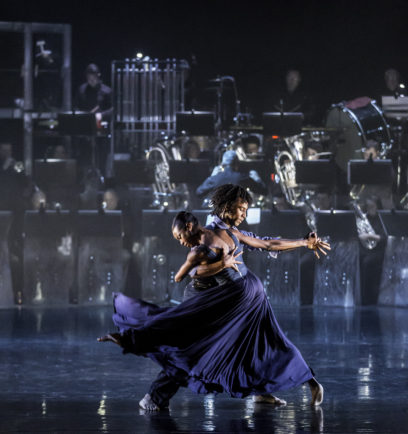Two dancers in flowing blue costumes perform a dramatic routine on stage, with a live orchestra playing in the background.