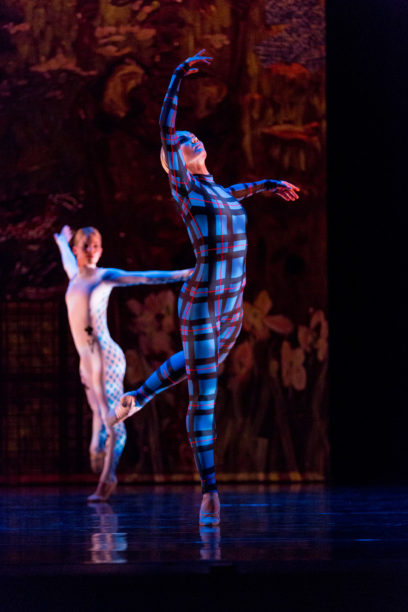 Two ballet dancers perform on stage, one in a blue and red checkered bodysuit and the other in a white and blue checkered bodysuit, against a colorful backdrop.
