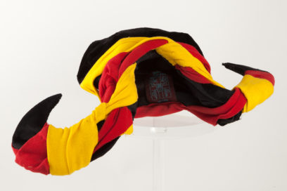 A multicolored jester's hat with curved, pointed tips in black, red, and yellow material, resting on a clear stand against a white background.