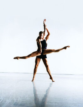 Two ballet dancers perform a lift in a well-lit studio, with one dancer extending their legs horizontally while being held in the air by the other dancer.