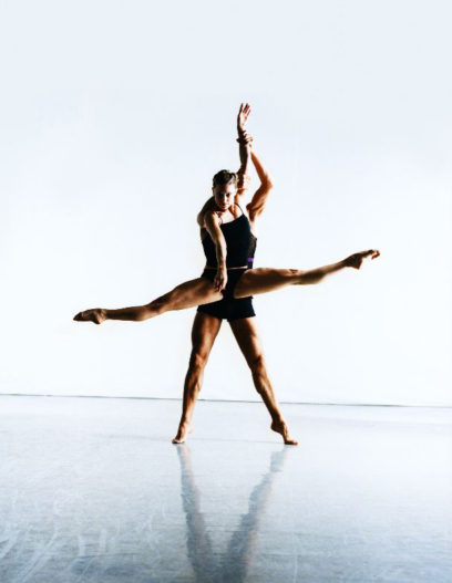 Two ballet dancers perform a lift in a well-lit studio, with one dancer extending their legs horizontally while being held in the air by the other dancer.