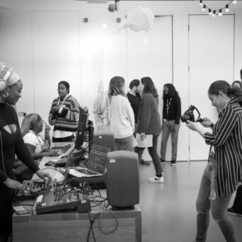 A group of people interact and work with various equipment in a room. A woman in the foreground operates DJ equipment, while another takes photos with a camera. Others engage in conversation in the background.