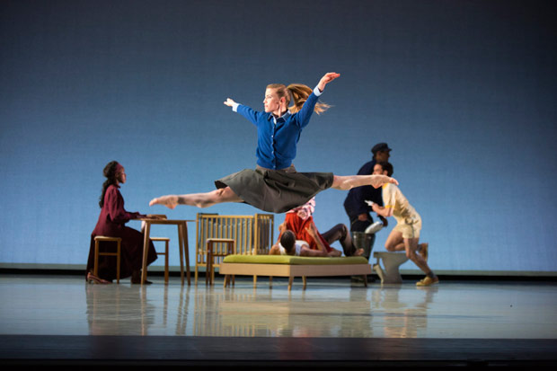 A dancer in a blue sweater and gray skirt performs a split jump on stage while other performers engage in various activities in the background.