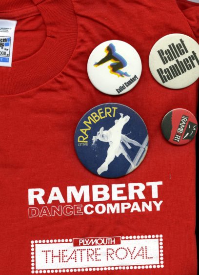 A red t-shirt with "Rambert Dance Company" and "Plymouth Theatre Royal" printed on it. Four badges are pinned to the shirt, each with designs related to Rambert and ballet.