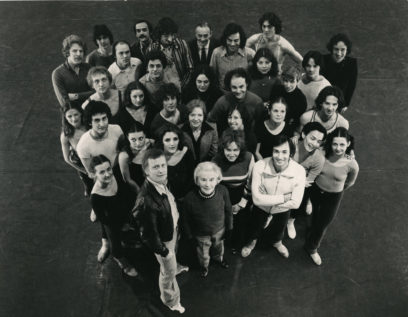 A group of 34 individuals, including men and women of varying ages, stand closely together in a formation with heads looking up at the camera. The photo is in black and white.