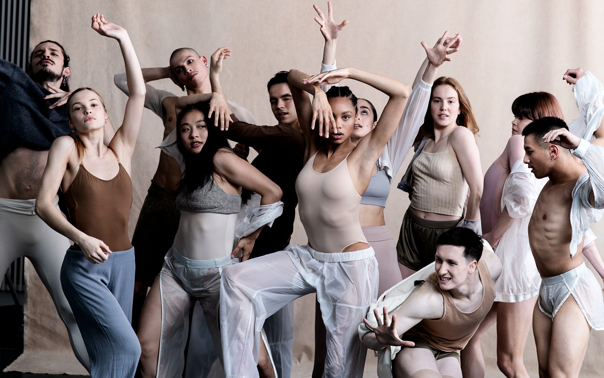 A diverse group of dancers in modern, neutral-toned attire strikes expressive poses against a plain backdrop.