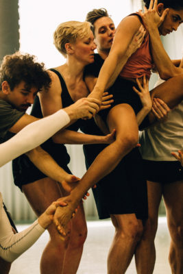 A group of people are collaborating in a dance, supporting one dancer who is elevated and reaching upwards.