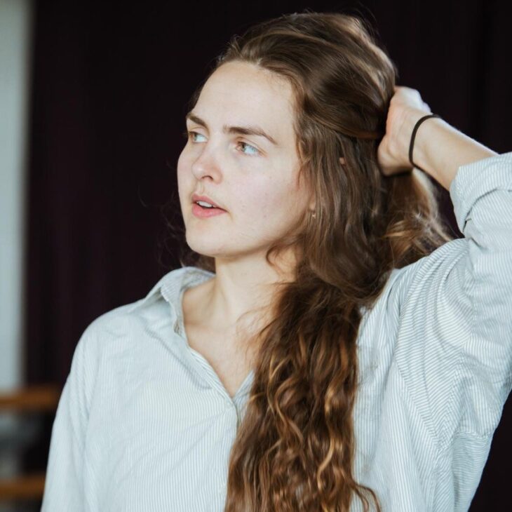 A person with long wavy hair and a light shirt stands against a dark background, looking to the side with one hand touching their hair.