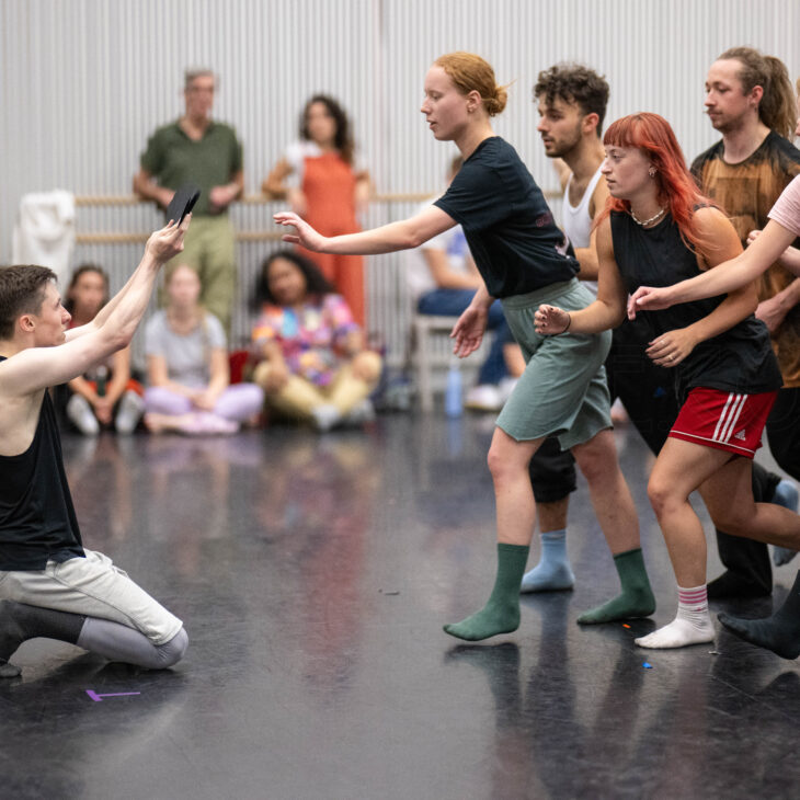 A group of people, some in casual athletic wear, reach out towards a kneeling individual in a dance studio, with onlookers in the background.