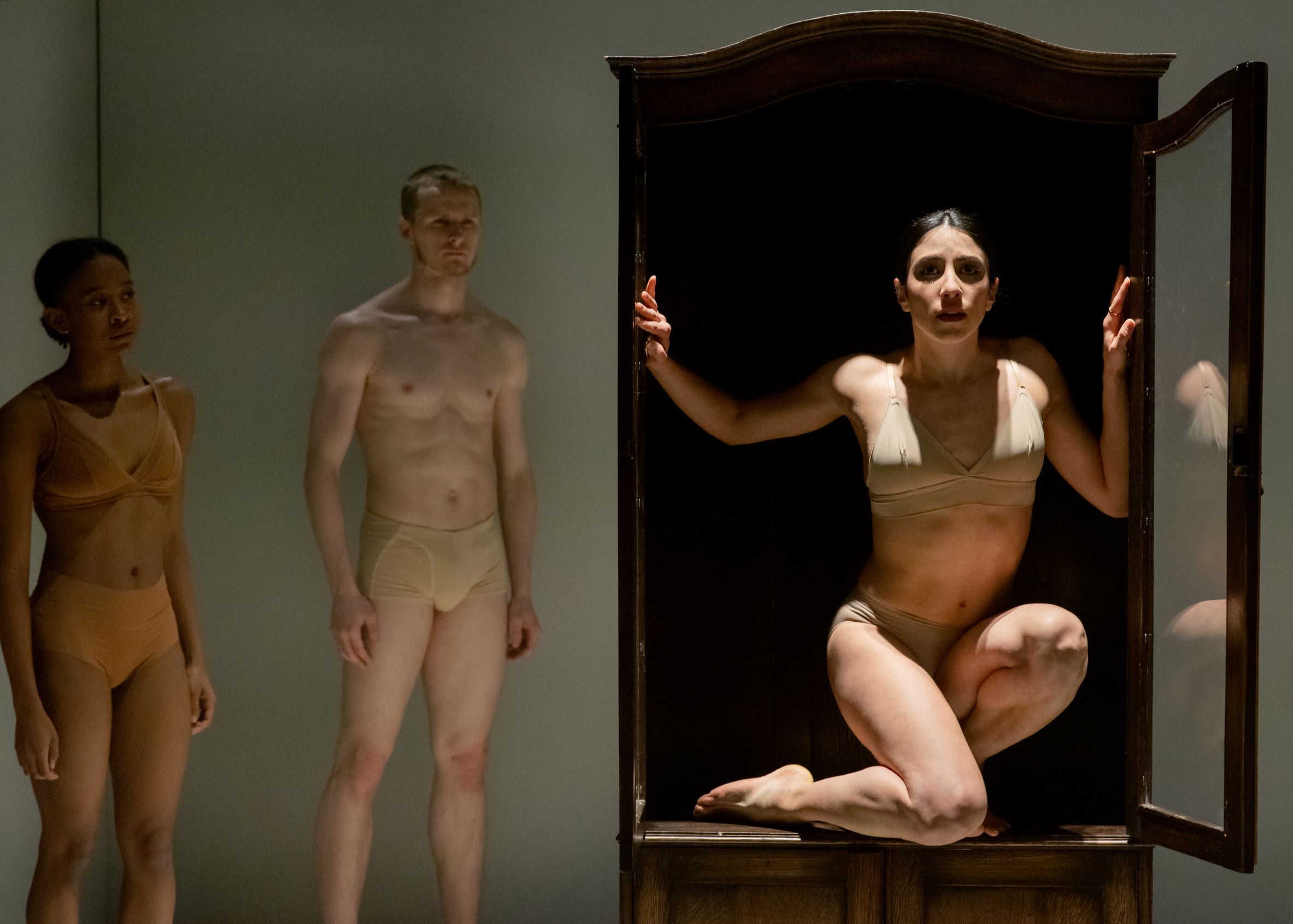 Three people in minimal clothing appear in a performance setting. One person stands inside a wardrobe with the door open, while the other two stand nearby, observing.