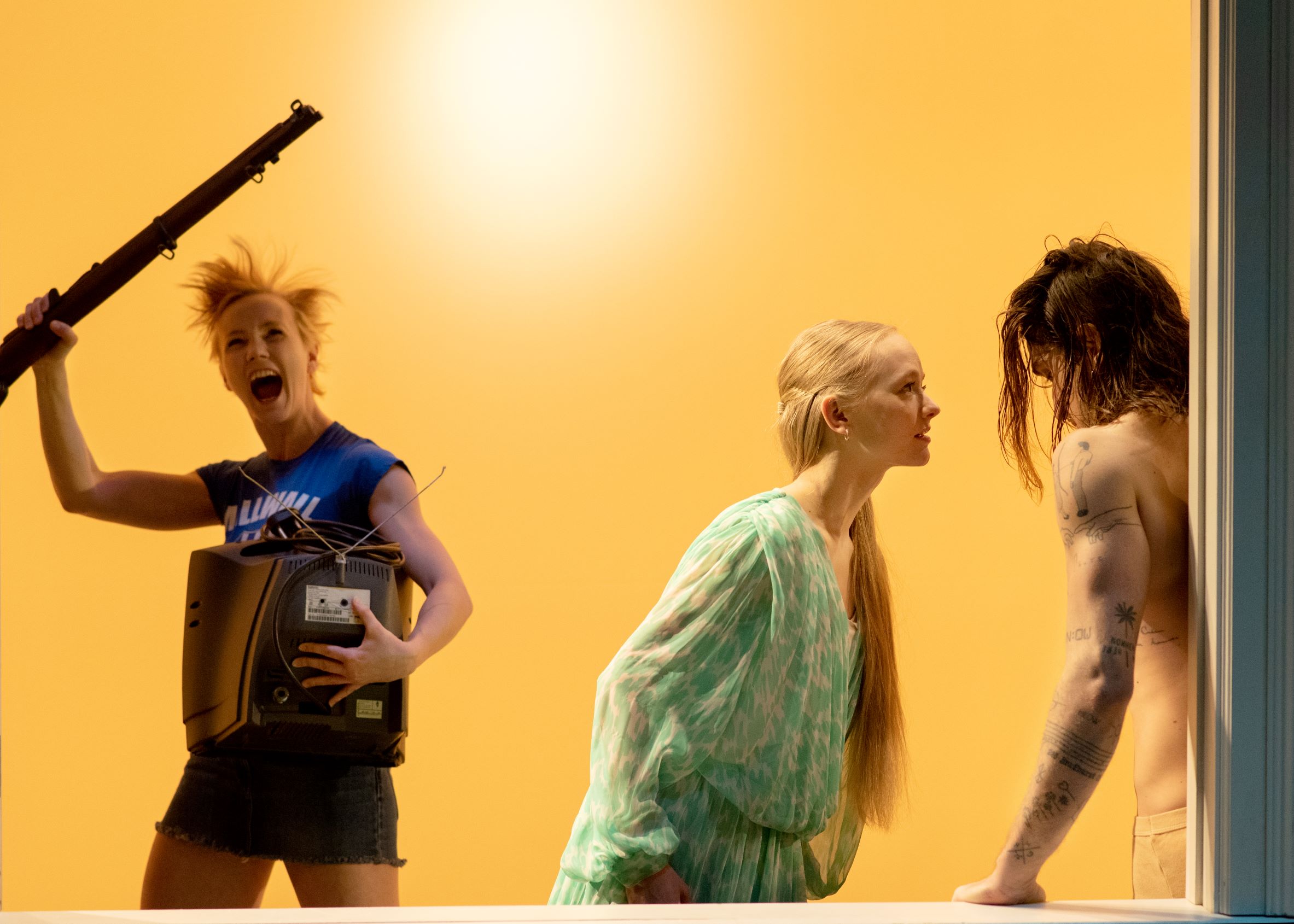 Three individuals in a brightly lit room; one holds a TV and a rifle, another with long hair is shirtless, and a third, dressed in green, leans forward.