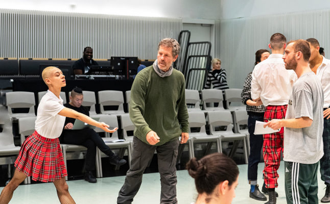A group of ballet dancers, including a bald dancer in a red plaid skirt, practice in a studio. An instructor in a green sweater oversees the rehearsal. Other dancers and staff are in the background.