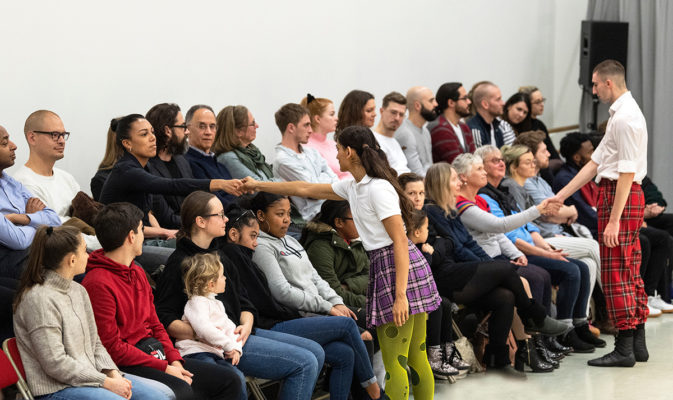 A group of people sits in rows facing performers who are interacting with them by reaching out and shaking hands. The audience includes children and adults dressed casually.