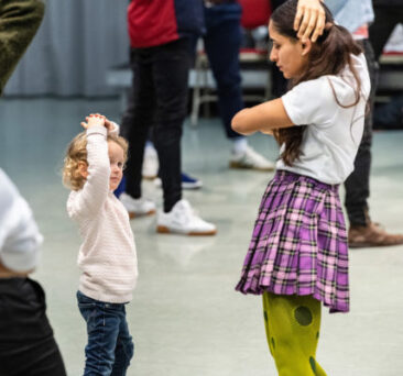 A young child and an adult stand facing each other indoors, both with their hands on their heads, imitating a similar pose. The room has a greenish floor and other people are visible in the background.