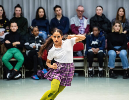 A dancer, in a white shirt, plaid skirt, and bright yellow tights, performs in front of a seated audience.