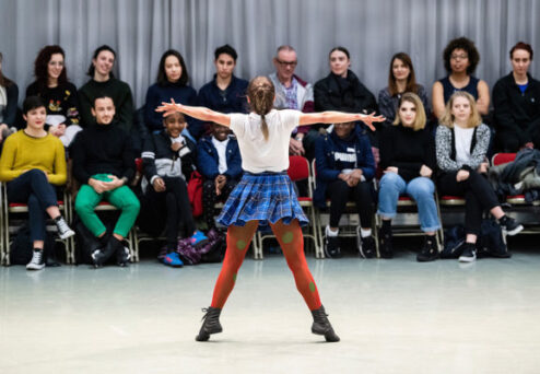 A person performs a dance in front of a seated audience. The performer is wearing a white shirt, a blue plaid skirt, red tights, and black shoes. The audience watches attentively.
