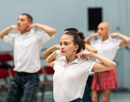 A group of dancers in white shirts and plaid skirts perform a routine with hands positioned near their necks, focused and synchronized.