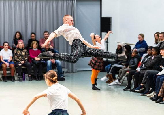 A dancer in a white shirt and plaid pants leaps mid-air while several dancers perform, and an audience watches in the background.