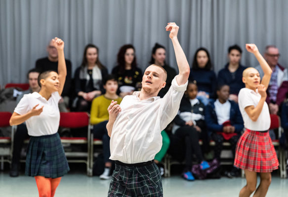 Three dancers in white shirts and plaid skirts perform in front of a seated audience. The background features a gray curtain.