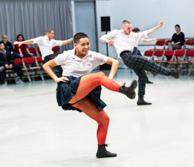 Two dancers wearing plaid skirts and white shirts perform high kicks on stage while a small audience watches in the background.