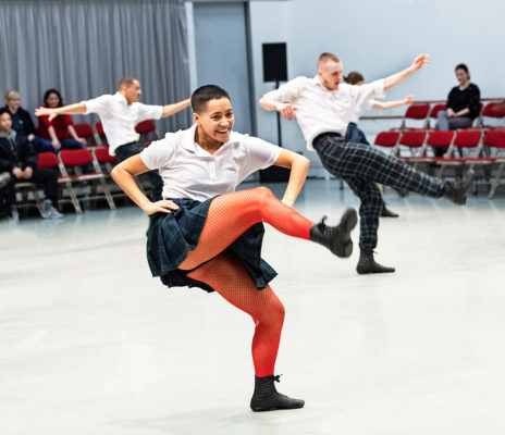 Two dancers wearing plaid skirts and white shirts perform high kicks on stage while a small audience watches in the background.