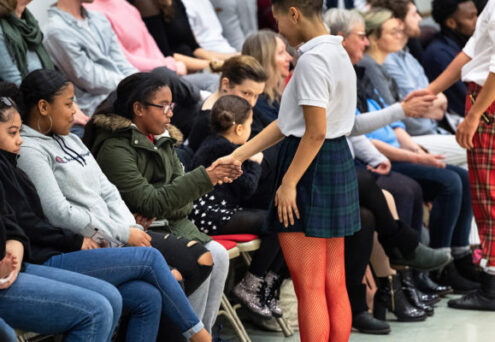 Person in a white shirt and checkered skirt shakes hands with an audience member. People sit on chairs in the background, watching attentively.