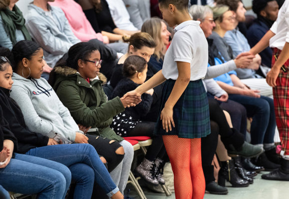 Person in a white shirt and checkered skirt shakes hands with an audience member. People sit on chairs in the background, watching attentively.