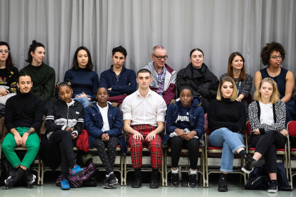 A group of people of varying ages sit in two rows of chairs against a gray curtain, looking towards the camera.