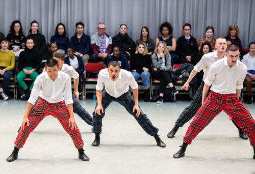 Five dancers in white shirts and plaid pants perform in front of a seated audience.
