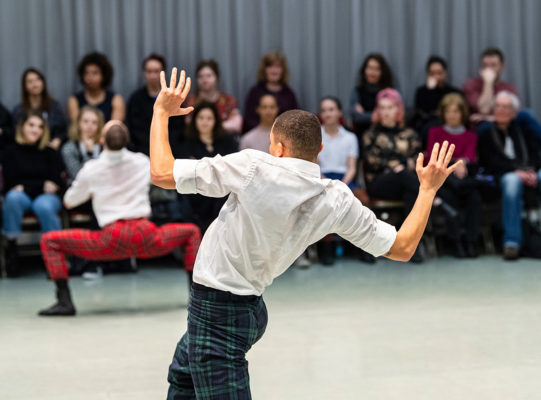 Two dancers in white shirts and plaid pants perform in front of a seated audience. One dancer appears to be crouching in the background, while the other stands with hands raised.