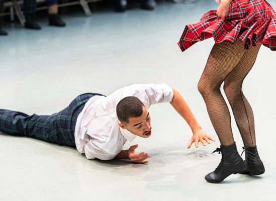 A person in a white shirt and plaid pants lies on the ground, reaching out towards another person in a plaid skirt and fishnet tights mid-dance.