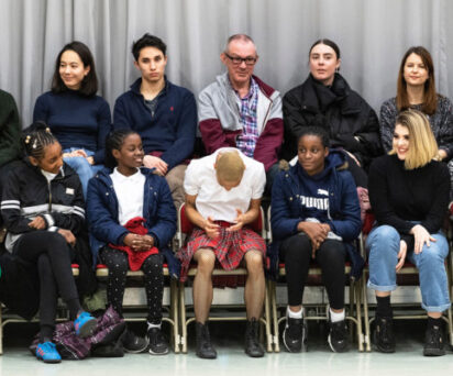 A group of people sitting in rows on chairs, some smiling and others looking ahead. A person in the front row, wearing a red plaid skirt, is looking down. A gray curtain is in the background.