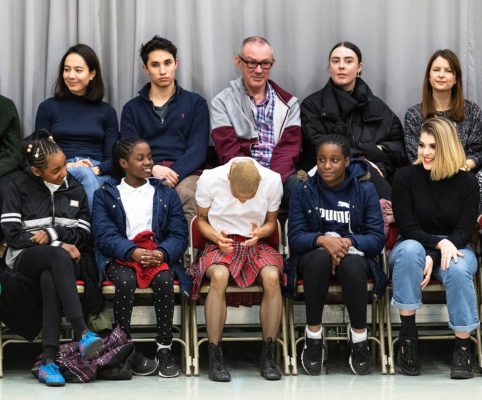A group of people sitting in rows on chairs, some smiling and others looking ahead. A person in the front row, wearing a red plaid skirt, is looking down. A gray curtain is in the background.