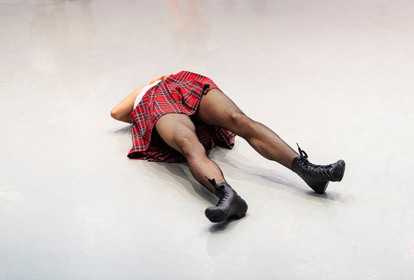 Person wearing a red plaid skirt, fishnet stockings, and black shoes lying face down on the floor.