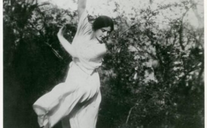 A person wearing flowy clothing is captured in an expressive dance pose outdoors, with one arm raised and the other extended to the side, against a background of trees and foliage.
