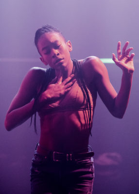 A person with braided hair and a focused expression performs a dance, one hand on their chest and the other raised. The lighting is soft and purple-toned.