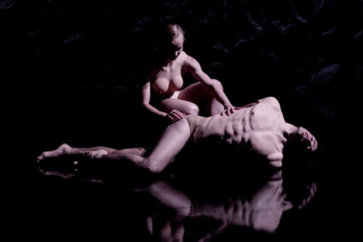 Two nude individuals in a dark, reflective environment; one is seated and touching the other's shoulder, who is lying down with an arched back.