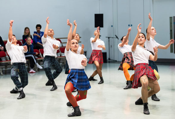 A group of dancers performs a choreographed routine wearing skirts and white tops, while an audience seated in the background watches.