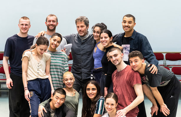 A group of thirteen people, including men and women, standing in a well-lit room with some of them hugging and smiling, with chairs lined up against a light blue wall in the background.