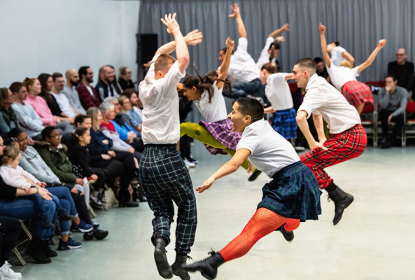 A group of dancers in colorful tartan skirts and white shirts leap into the air in front of a seated audience in a performance space.