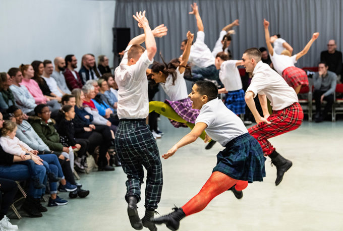 Dancers in colorful tartan skirts and white shirts perform an energetic leap in a room with seated audience members watching.