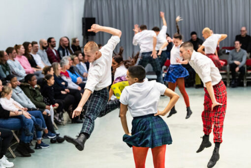 Performers in plaid skirts and white shirts, mid-jump during a dance routine. An audience sits in rows of chairs, attentively watching the performance in a brightly lit room.