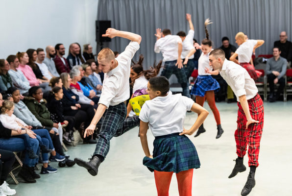 Performers in plaid skirts and white shirts, mid-jump during a dance routine. An audience sits in rows of chairs, attentively watching the performance in a brightly lit room.