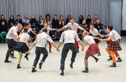 A group of people in kilts and white shirts dance in a circle while holding hands. An audience sits in the background watching the performance.