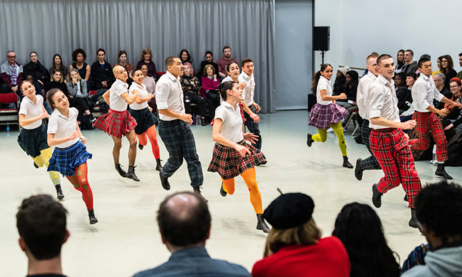 A dance troupe, wearing plaid skirts and colorful tights, performs energetically on stage in front of a seated audience.