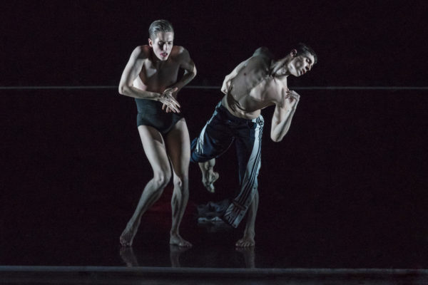 Two shirtless dancers perform on a dark stage, one wearing dark shorts and the other in striped pants, both in dynamic, expressive poses.