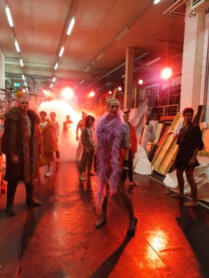 A group of people in costumes stands in a dimly lit area with a red light illuminating the background. One person in the center is wearing a feather boa. Various props and set pieces are scattered around.