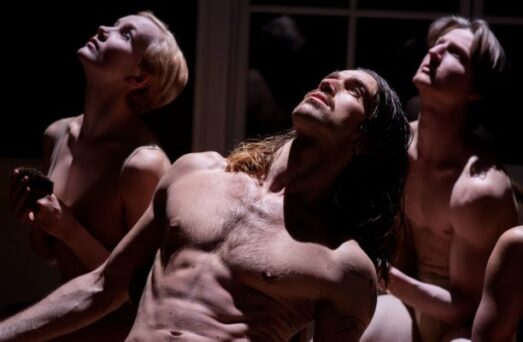 Three shirtless individuals stand closely together in a dark setting, looking up intensely towards a light source.