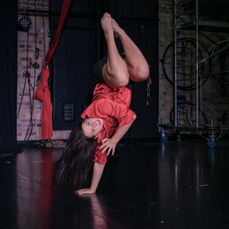 A dancer who has long brown hair and is wearing a red long sleeve shirt that stops at their elbows and black shorts. They are in a balance upside down on one arm with their legs bent into their chest and is looking straight forward.