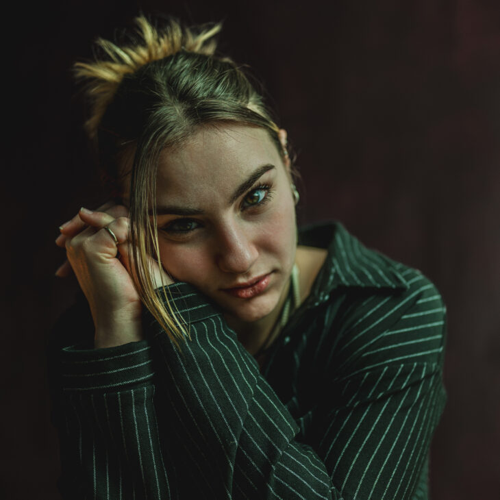 A dancer who has blonde hair tied back and is wearing a dark green striped long-sleeve shirt. They are leaning onto their hands which are clasped together and they are looking straight forwards.